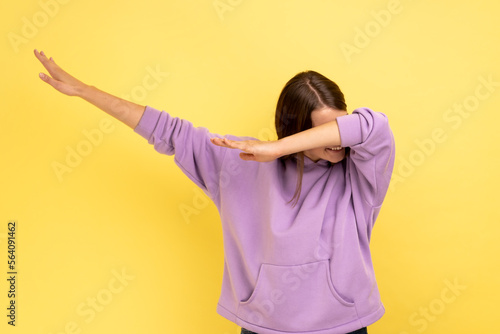 Winning, success gesture. Portrait of woman showing dab dance pose, famous internet meme, performing dabbing trends, wearing purple hoodie. Indoor studio shot isolated on yellow background.