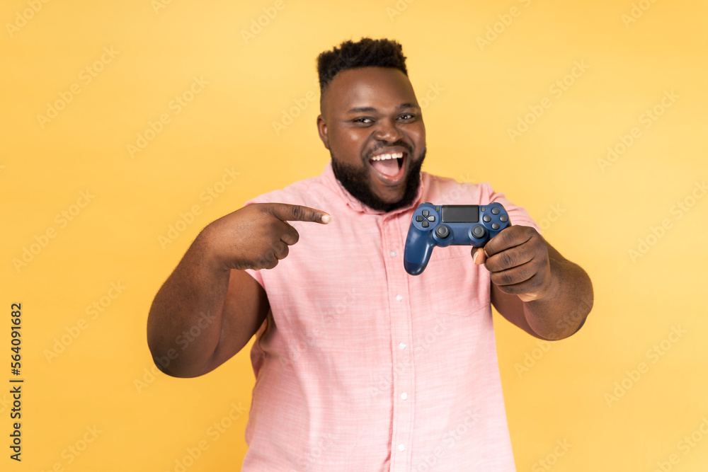 Portrait of excited happy positive bearded man wearing pink shirt standing with excited expression, holding pointing at joystick. Indoor studio shot isolated on yellow background. Portrait of excited happy positive bearded man wearing pink shirt standing with excited expression, holding pointing at joystick. Indoor studio shot isolated on yellow background.