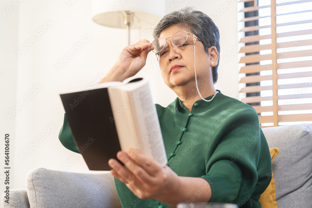 Presbyopia, Hyperopia mature asian woman holding eyeglasses having problem with vision problem trying to read text on book, eye disease of old elderly sitting on couch. Poor eyesight, health care. Presbyopia, Hyperopia mature asian woman holding eyeglasses having problem with vision problem trying to read text on book, eye disease of old elderly sitting on couch. Poor eyesight, health care.