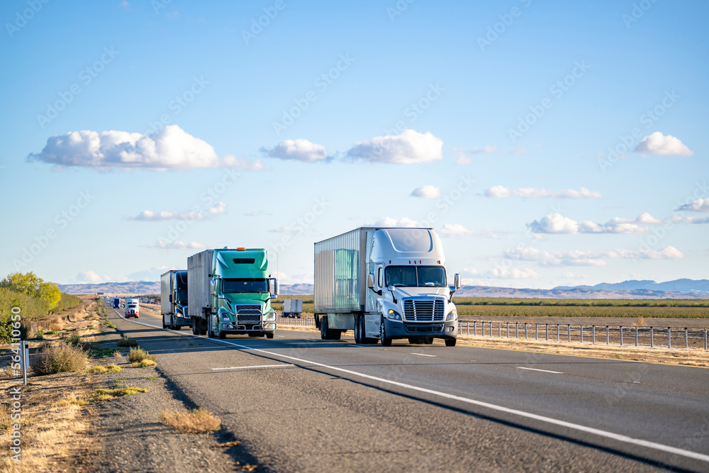 Convoy of the big rig semi trucks with different semi trailers carry cargo running on the divided highway road in both directions