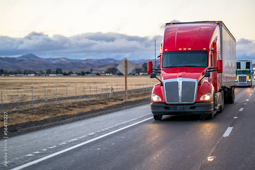 Heavy duty red big rig semi truck with dry van semi trailer running on the straight road in front of another semi truck at twilight time Heavy duty red big rig semi truck with dry van semi trailer running on the straight road in front of another semi truck at twilight time