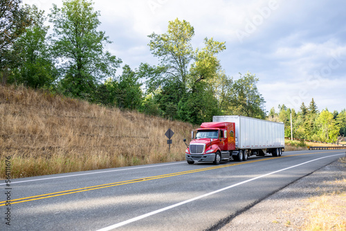 Low cab profile red big rig semi truck transporting goods in dry van semi trailer driving on the narrow mountain road