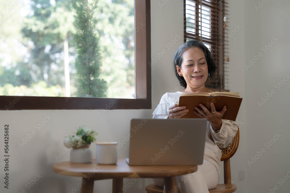 Relaxed and calm 60s retired Asian woman looking out the window, daydreaming while reading a book in her home living room. lifestyle concept Relaxed and calm 60s retired Asian woman looking out the window, daydreaming while reading a book in her home living room. lifestyle concept