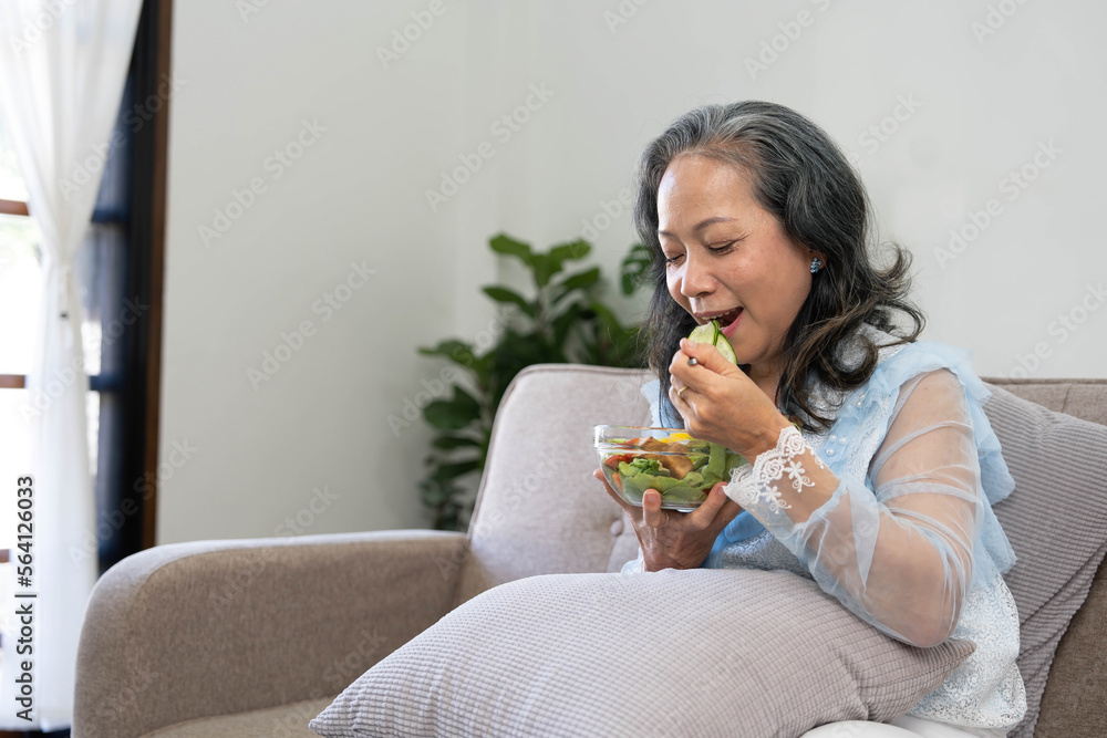 Portrait, Cheerful Asian 60s aged woman having breakfast in her living room, eating healthy salad vegetables mix bowl. Portrait, Cheerful Asian 60s aged woman having breakfast in her living room, eating healthy salad vegetables mix bowl.