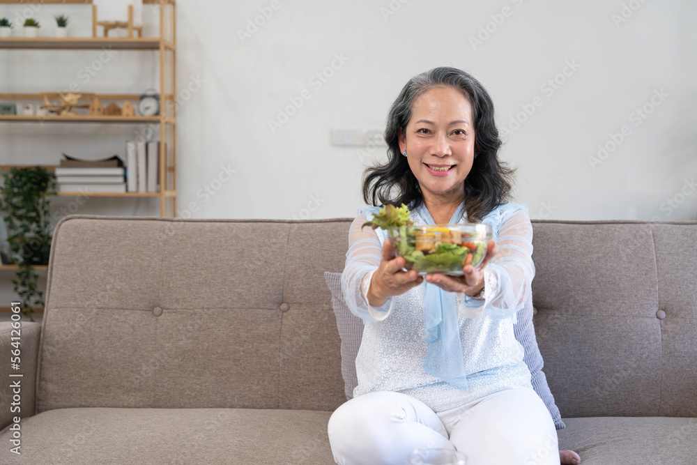 Portrait, Cheerful Asian 60s aged woman having breakfast in her living room, eating healthy salad vegetables mix bowl. Portrait, Cheerful Asian 60s aged woman having breakfast in her living room, eating healthy salad vegetables mix bowl.
