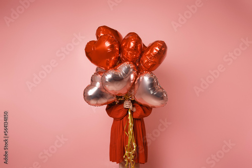 The girl is holding red and pink balloons in front of her on a pink background in a red dress. Young woman on birthday holiday party. Card post Valentine's Day, February 14.