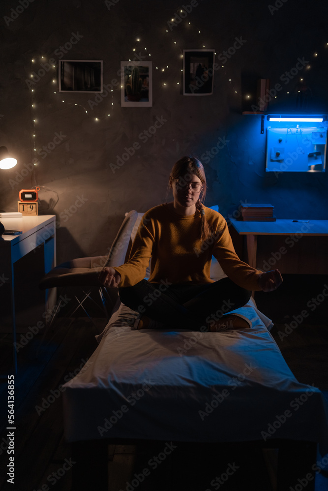 Young woman meditating on bed in lotus position sitting in dark room at night