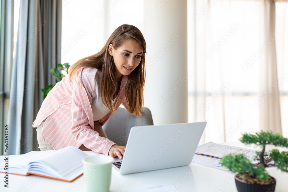 Beautiful smiling businesswoman reading something on a laptop while leaning on the table in the office Beautiful smiling businesswoman reading something on a laptop while leaning on the table in the office