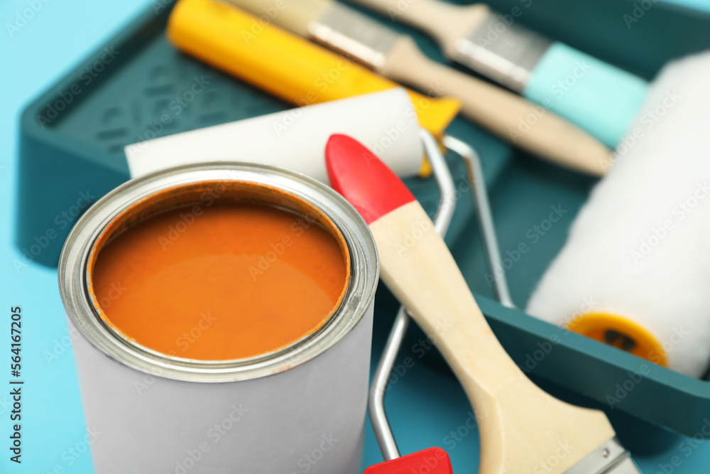 Can of orange paint, brushes, rollers and container on turquoise background, closeup