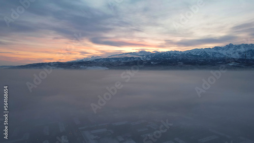 Dawn over the city in fog and smog. Mountain view. A light haze hangs over the city, cars drive, signs glow. Clouds, sun rays and snowy mountains are visible in the distance. Almaty, Kazakhstan