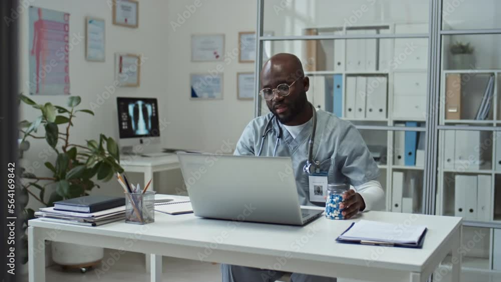 Young confident African American online doctor in uniform sitting by desk in front of laptop in clinics, giving medical advice to patient and recommending new effective medicament Young confident African American online doctor in uniform sitting by desk in front of laptop in clinics, giving medical advice to patient and recommending new effective medicament