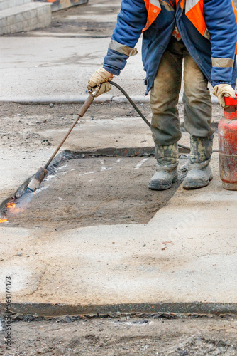 A worker repairs a road by heating a damaged asphalt pavement with a gas burner.