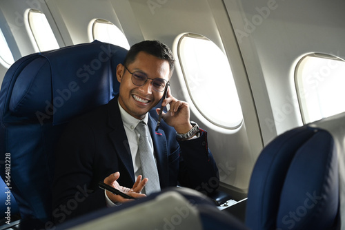 Confident businessman in black suit sitting comfortable seat in airplane cabin and talking on mobile phone © Prathankarnpap