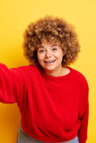Close up of beautiful smiling charming young curly haired woman 30s wears red sweater, doing selfie on mobile phone, isolated on yellow background studio portrait. People emotions lifestyle concept