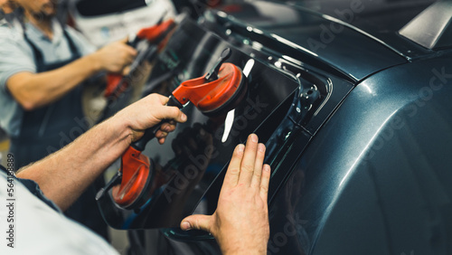 Close-up of two unrecognisable men holding rear window pane to install into back of car. Car maintenance. Garage work. Horizontal indoor shot. High quality photo