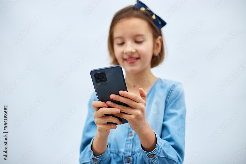 Smiling little girl in casual denim dress hold smartphone, using mobile cell phone internet applications, typing message isolated on white background, studio. Child and modern tech usage concept Smiling little girl in casual denim dress hold smartphone, using mobile cell phone internet applications, typing message isolated on white background, studio. Child and modern tech usage concept