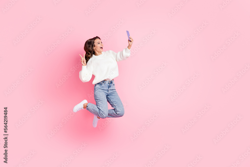 Full length photo of friendly woman dressed pullover jumping showing v-sign device empty space isolated pink color background