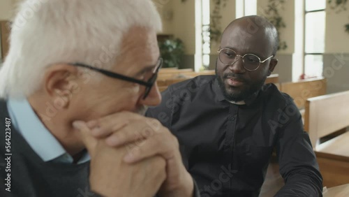Selective focus shot of senior Caucasian man attending Catholic church speaking about his problems, young adult Black priest comforting himg