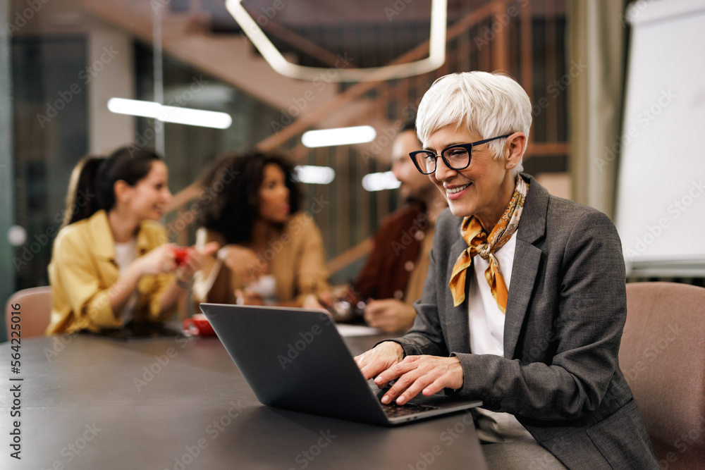 Smiling businesswoman working over the laptop, being in the office with her colleagues. Smiling businesswoman working over the laptop, being in the office with her colleagues.