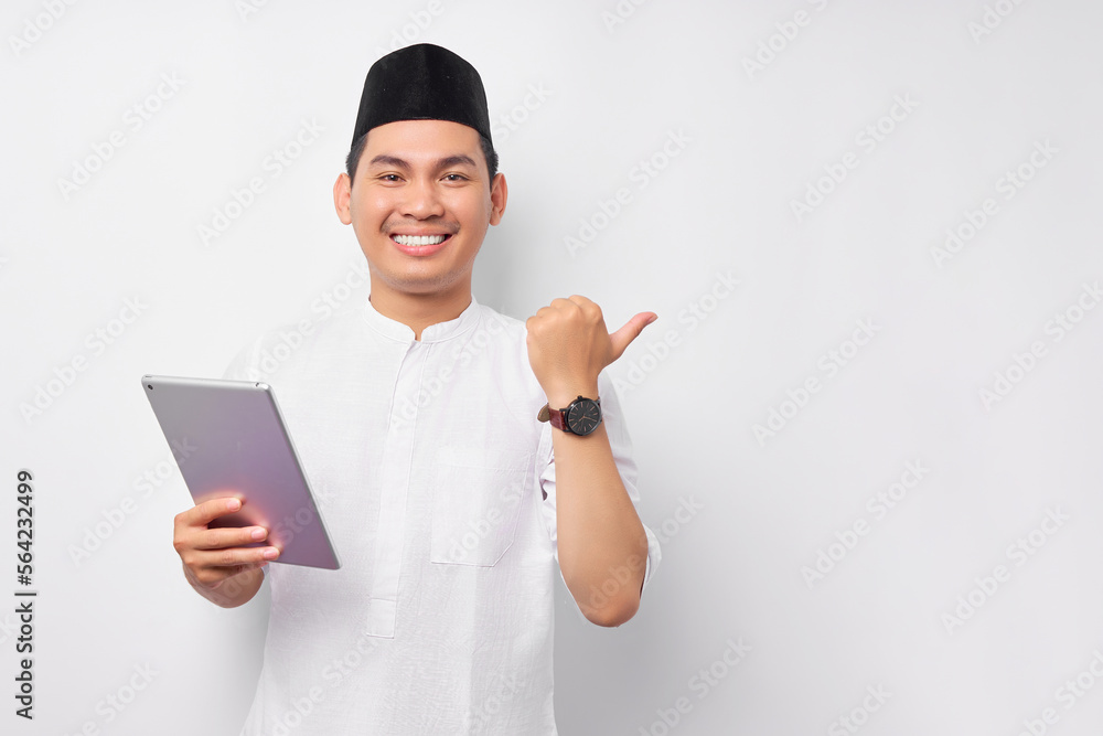 Cheerful young Asian Muslim man holding digital tablet and pointing thumb at copy space isolated on white background. People religious Islam lifestyle concept. celebration Ramadan and ied Mubarak Cheerful young Asian Muslim man holding digital tablet and pointing thumb at copy space isolated on white background. People religious Islam lifestyle concept. celebration Ramadan and ied Mubarak