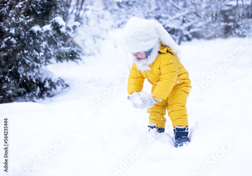 child playing in snow. cheerful little girl in yellow snowsuit and fluffy white hat having fun with snow on winter day in the park