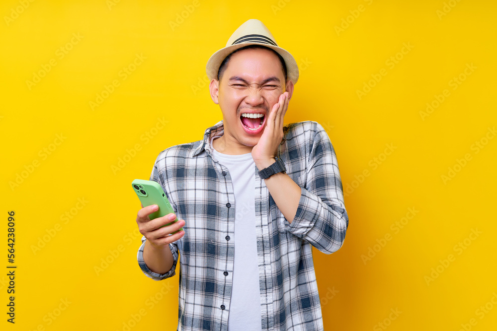 funny Young handsome ethnic Asian man 20s wearing casual clothes hat holding mobile phone with his hand touching his cheek isolated on yellow background. People lifestyle concept