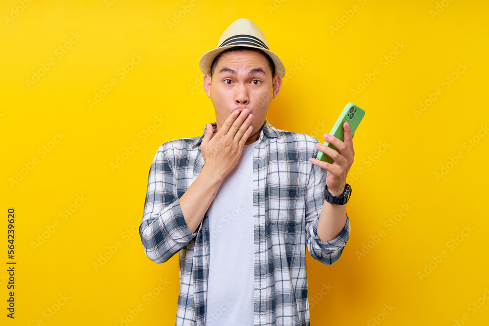 Surprised handsome Asian young man 20s wearing casual clothes hat cover mouth with hand look camera while holding mobile phone isolated on yellow background. People lifestyle concept