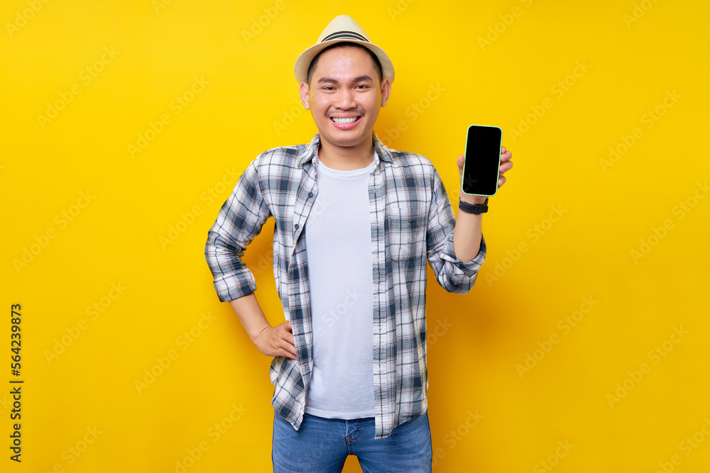 Cheerful handsome Asian young man 20s wearing casual clothes hat standing while hand on his waist, Showing mobile cell phone with blank screen isolated on yellow background. People lifestyle concept