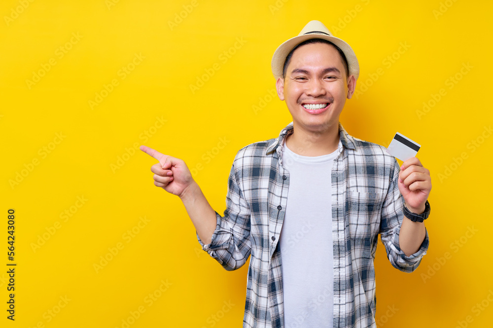 Smiling happy Asian ethnicity young man 20s wearing casual clothes hat holding in hand credit bank card, pointing finger aside isolated on yellow background studio portrait. People lifestyle concept Smiling happy Asian ethnicity young man 20s wearing casual clothes hat holding in hand credit bank card, pointing finger aside isolated on yellow background studio portrait. People lifestyle concept
