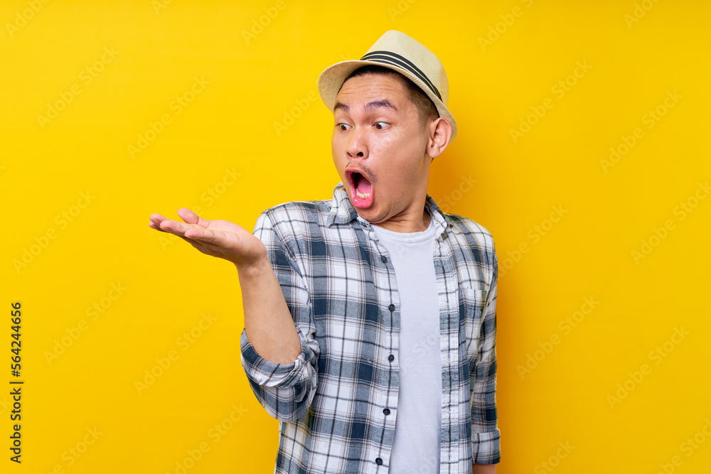 Surprised Asian ethnicity young man 20s wearing casual clothes hat looking palm, imagine the product isolated on yellow background studio portrait. People lifestyle concept Surprised Asian ethnicity young man 20s wearing casual clothes hat looking palm, imagine the product isolated on yellow background studio portrait. People lifestyle concept