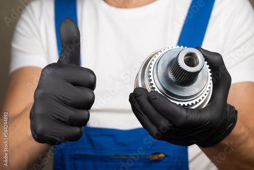 Murais de parede Auto mechanic in blue jumpsuit holds in his hands hinge of equal angular velocities of car and rotates it in black gloves