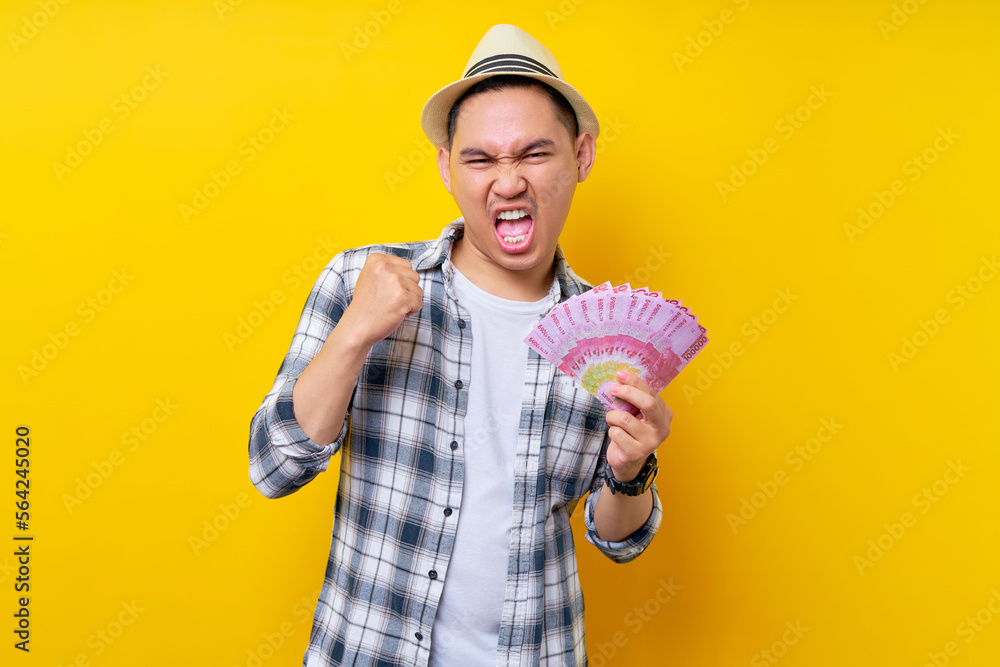 Excited Young handsome ethnic Asian man 20s wearing casual clothes hat holding cash in rupiah banknotes, celebrating luck isolated on yellow background. People lifestyle concept Excited Young handsome ethnic Asian man 20s wearing casual clothes hat holding cash in rupiah banknotes, celebrating luck isolated on yellow background. People lifestyle concept