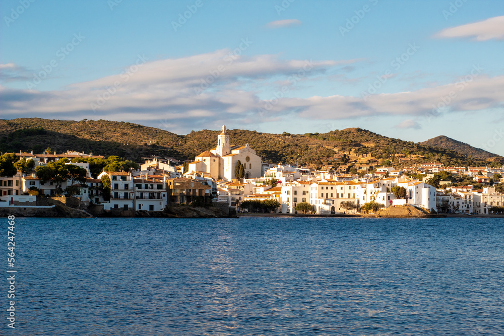 Panorámica del blanco pueblo pesquero Cadaqués en la Costa Brava con las verdes montañas de fondo y el traqnuilo mar Mediterráneo por delante.