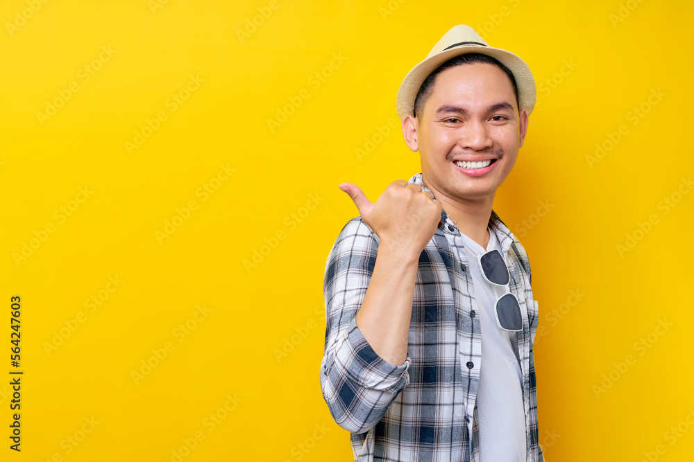 Smiling cheerful Young handsome ethnic Asian man 20s wearing casual clothes hat standing pointing thumb aside on mock up copy space isolated on yellow background. People lifestyle concept