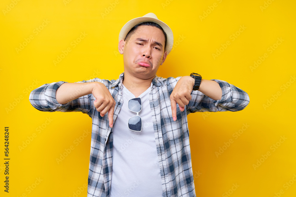 Sadness Young handsome ethnic Asian man 20s wearing casual clothes hat standing pointing index fingers down on mock up copy space isolated on yellow background. People lifestyle concept