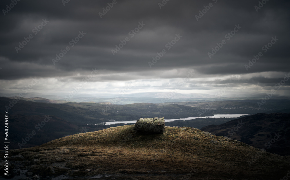 A large boulder on top of Raven Crag overlooking Lake Windermere in the distance in the Cumbrian Lake District Mountains in winter, England UK.