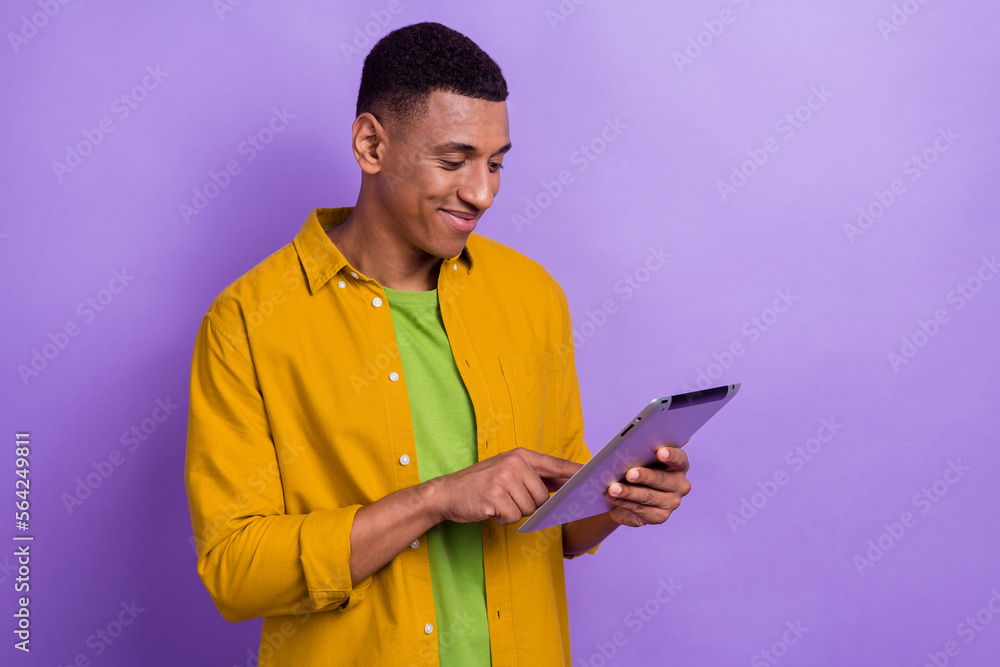 Photo portrait of nice young guy holding tablet gadget typing scrolling wear trendy yellow garment isolated on purple color background Photo portrait of nice young guy holding tablet gadget typing scrolling wear trendy yellow garment isolated on purple color background