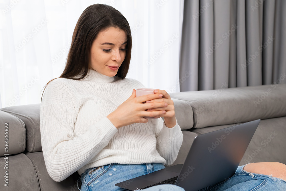 Beautiful brunette with long hair holding a cup watching video, having a pleasant conversation online using computer in cozy living room. Beautiful brunette with long hair holding a cup watching video, having a pleasant conversation online using computer in cozy living room.