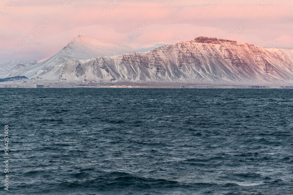 imagen de unas montañas iluminadas por las últimas luces del día, dando un tono magenta, con el cielo nublado y el mar en la parte inferior