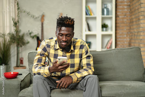 Handsome African man using smartphone while sitting on a sofa in cozy living room. Jpung man typing a message.