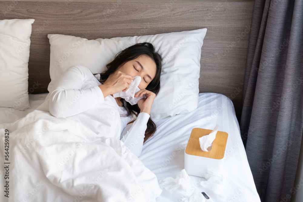 Young Asian woman sneezing, flu or allergy symptoms blowing nose on bed