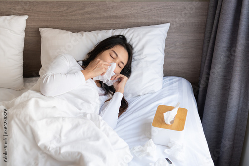 Young Asian woman sneezing, flu or allergy symptoms blowing nose on bed after wake up in the morning. Illness, home isolation  concept.