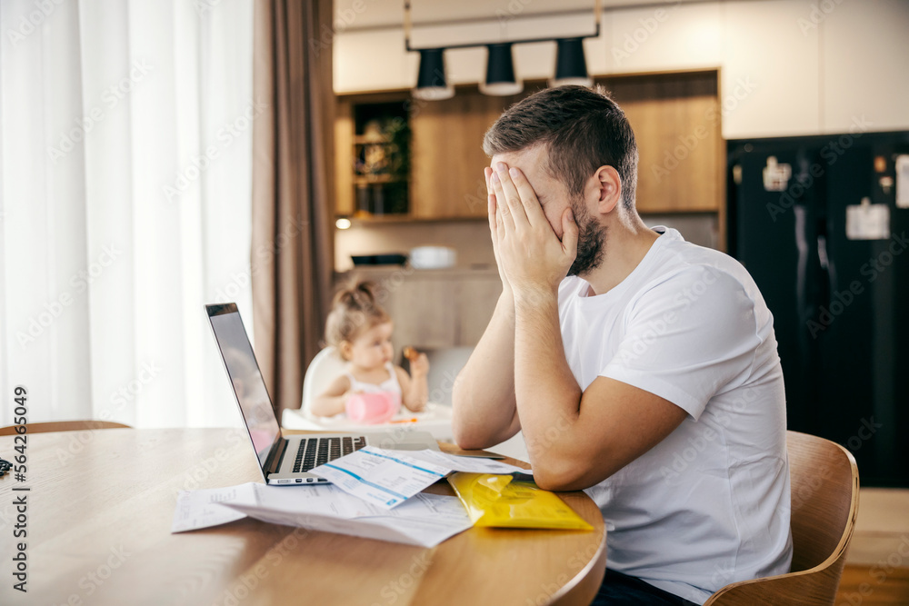 A disappointed man is sitting in front of his laptop and paying bills while his daughter is playing in blurry background.