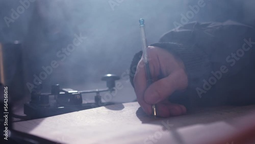 Man sends signals on telegraph by tapping finger on telegraph and makes notes in diary. Male person sits at table in dark room closeup photo
