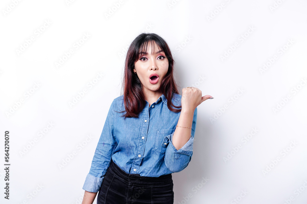 © Reezky - Shocked Asian woman wearing blue shirt pointing at the copy space beside her, isolated by white background © Reezky - Shocked Asian woman wearing blue shirt pointing at the copy space beside her, isolated by white background