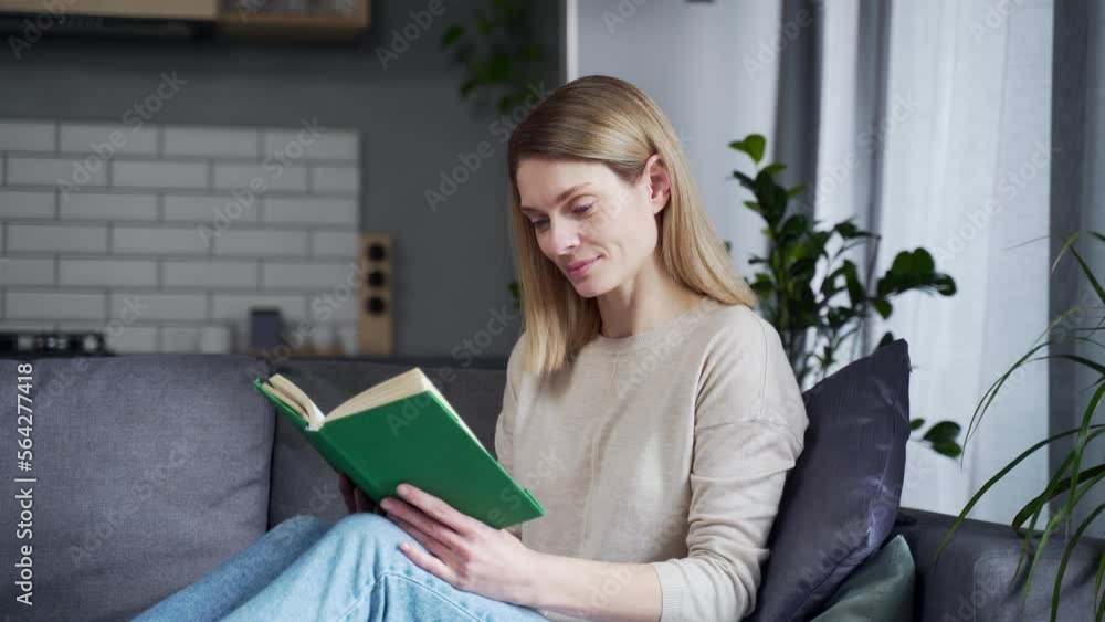 Close-up. Mature beautiful woman reading a book while sitting on the sofa in the living room at home. Relaxing on a comfortable couch during free time on weekends. Happy female student with a textbook Close-up. Mature beautiful woman reading a book while sitting on the sofa in the living room at home. Relaxing on a comfortable couch during free time on weekends. Happy female student with a textbook
