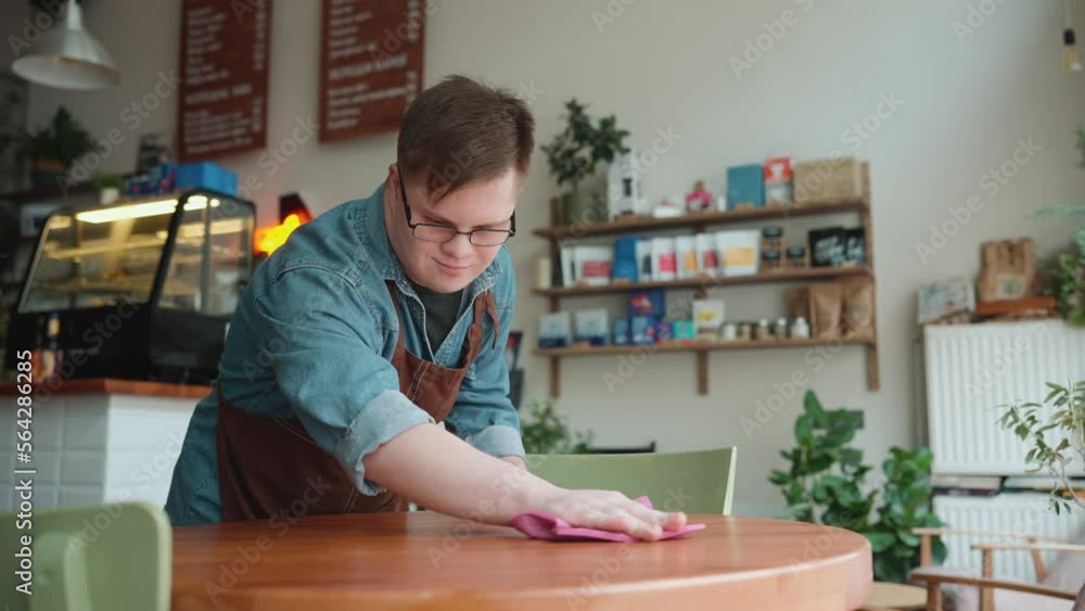Positive man barista with Down syndrome wiping table in cafe Positive man barista with Down syndrome wiping table in cafe
