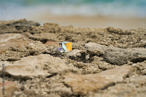 Fototapeta Naklejka Na Ścianę i Meble -  Geschliffenes, bunt schimmerndes Glas Quadrat auf einem Felsen am Strand, abstrakt und anders