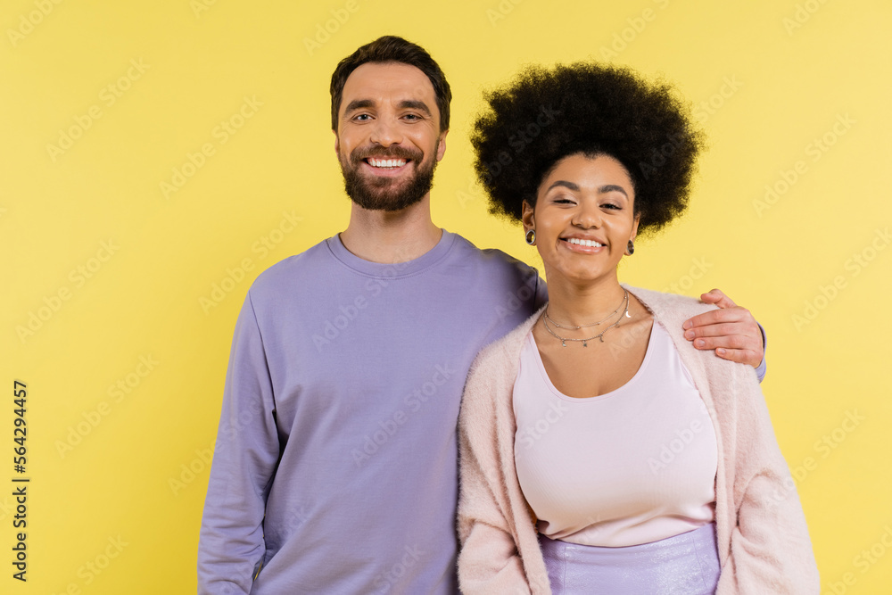 happy bearded man hugging shoulders of stylish african american woman while looking at camera isolated on yellow. happy bearded man hugging shoulders of stylish african american woman while looking at camera isolated on yellow.