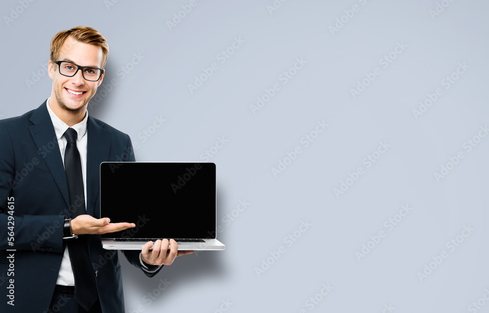 Business man in glasses spectacles, black suit showing laptop with empty mockup screen, on grey color background, with copy space. It expert,  technician repair service, tech support, maintenance.